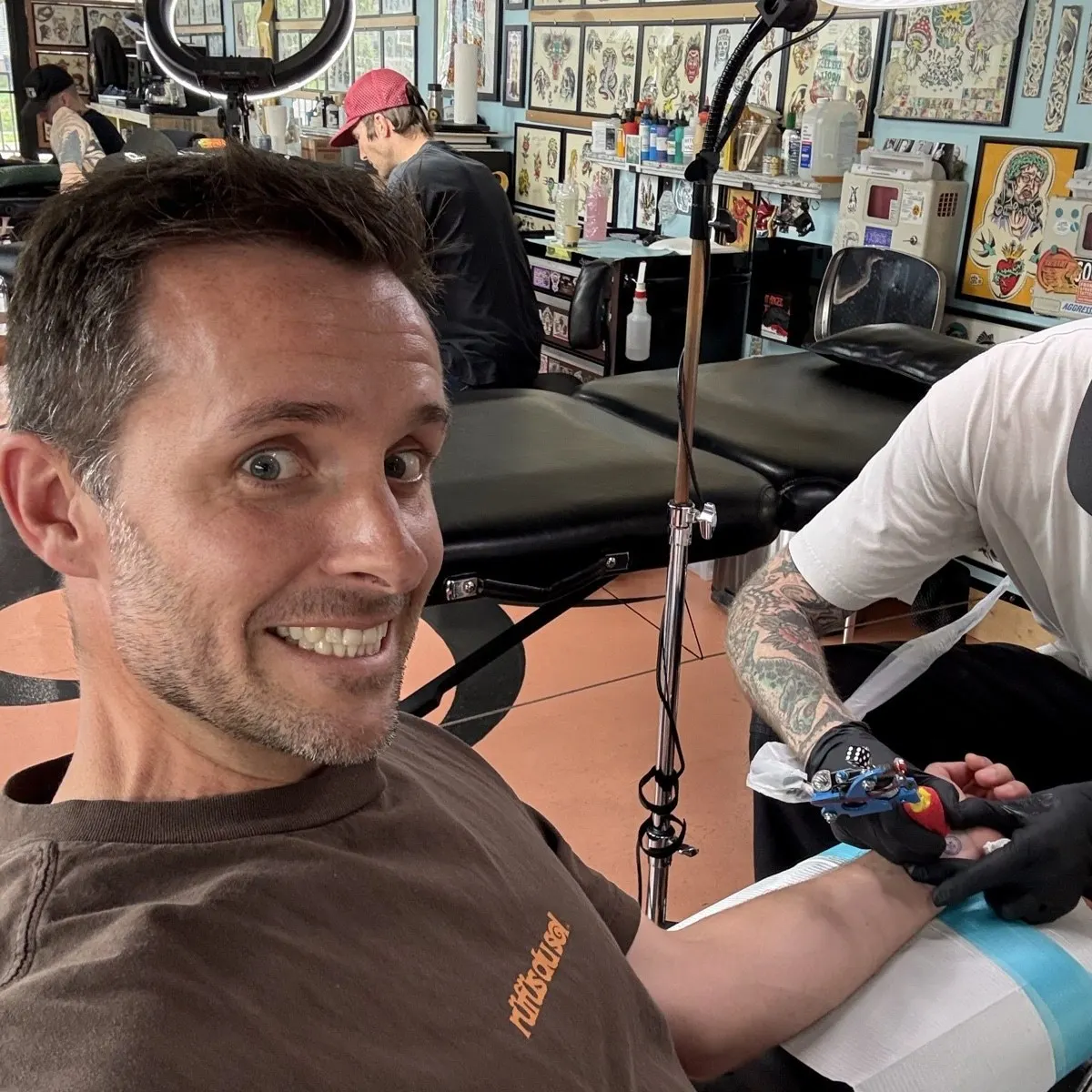 Ryan grinning in a tattoo shop while an artist works on his arm